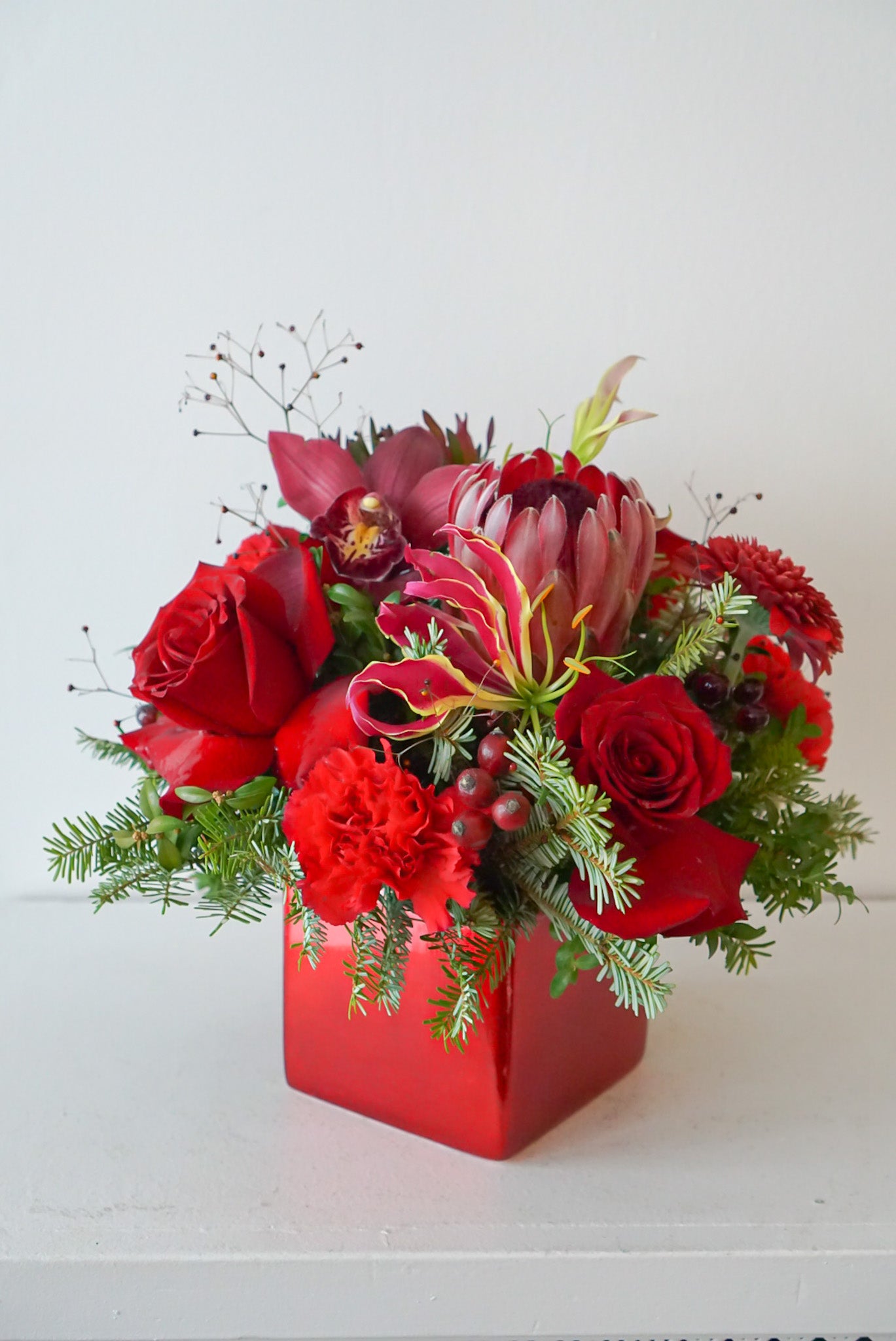 Bring your holiday table to life with this beautiful all red blooms flower arrangement. A charming display of protea, freedom roses, gloriosa, with a fragrant assortment of winter evergreen. Flower Nook, Toronto Florist