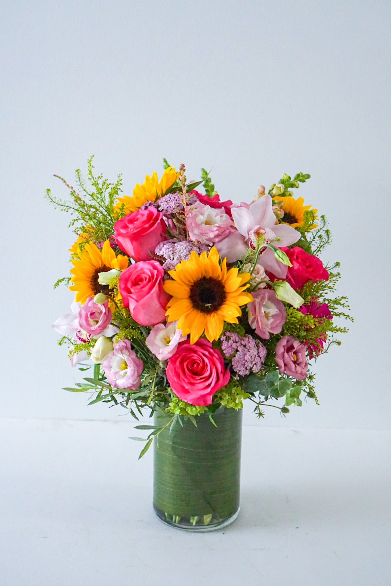 A bright summery arrangement, Summer Soiree is an ideal gift sure to brighten their day. This vivacious bouquet is an assortment of hot pink, yellow, orange and green summer blooms. Delivered in a clear cylinder vase, lined with green ti leave Sunflowers, roses, lisianthus, yallow, cymbidium orchid, snap dragon, solidago, green hydrangeas. The Flower Nook- Toronto Florist- Flowers delivery Toronto