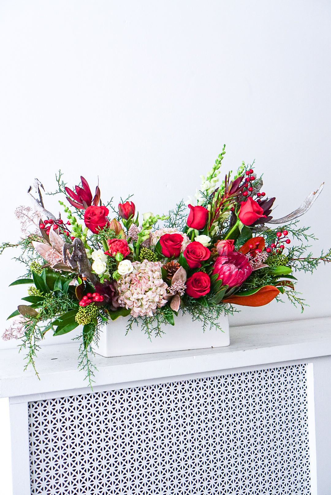 Holiday centerpiece in white rectangular vase with protea, red roses, mums, and evergreens by The Flower Nook