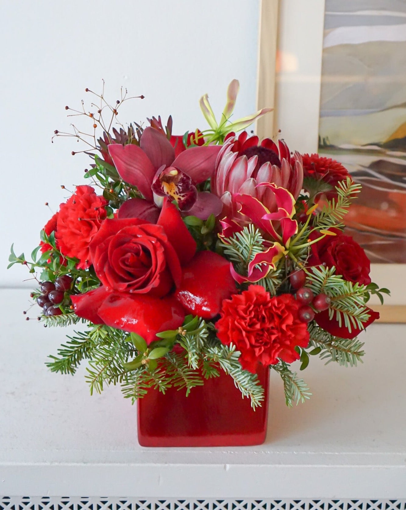 Bring your holiday table to life with this beautiful all red blooms flower arrangement. A charming display of protea, freedom roses, gloriosa, with a fragrant assortment of winter evergreen. Flower Nook, Toronto Florist