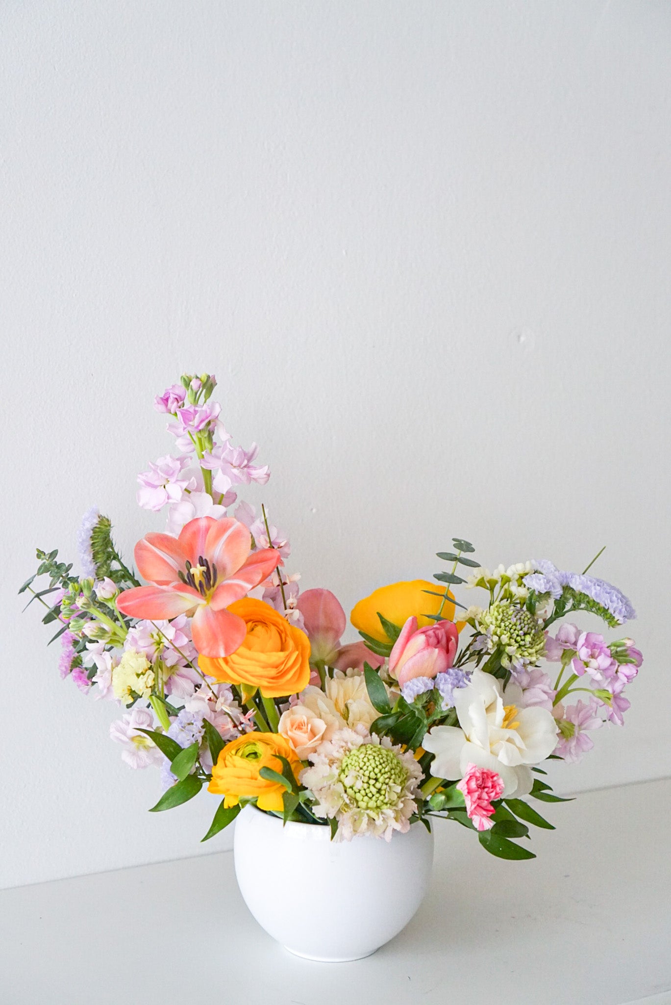 Like a spectacular spring sunrise, this radiant arrangement of yellow, peach, pink blooms, designed in a modern circular pot. This arrangement is sure to brighten anyone's day!  This arrangement features ranunculus, tulips, stocks, scabiosa, statics.