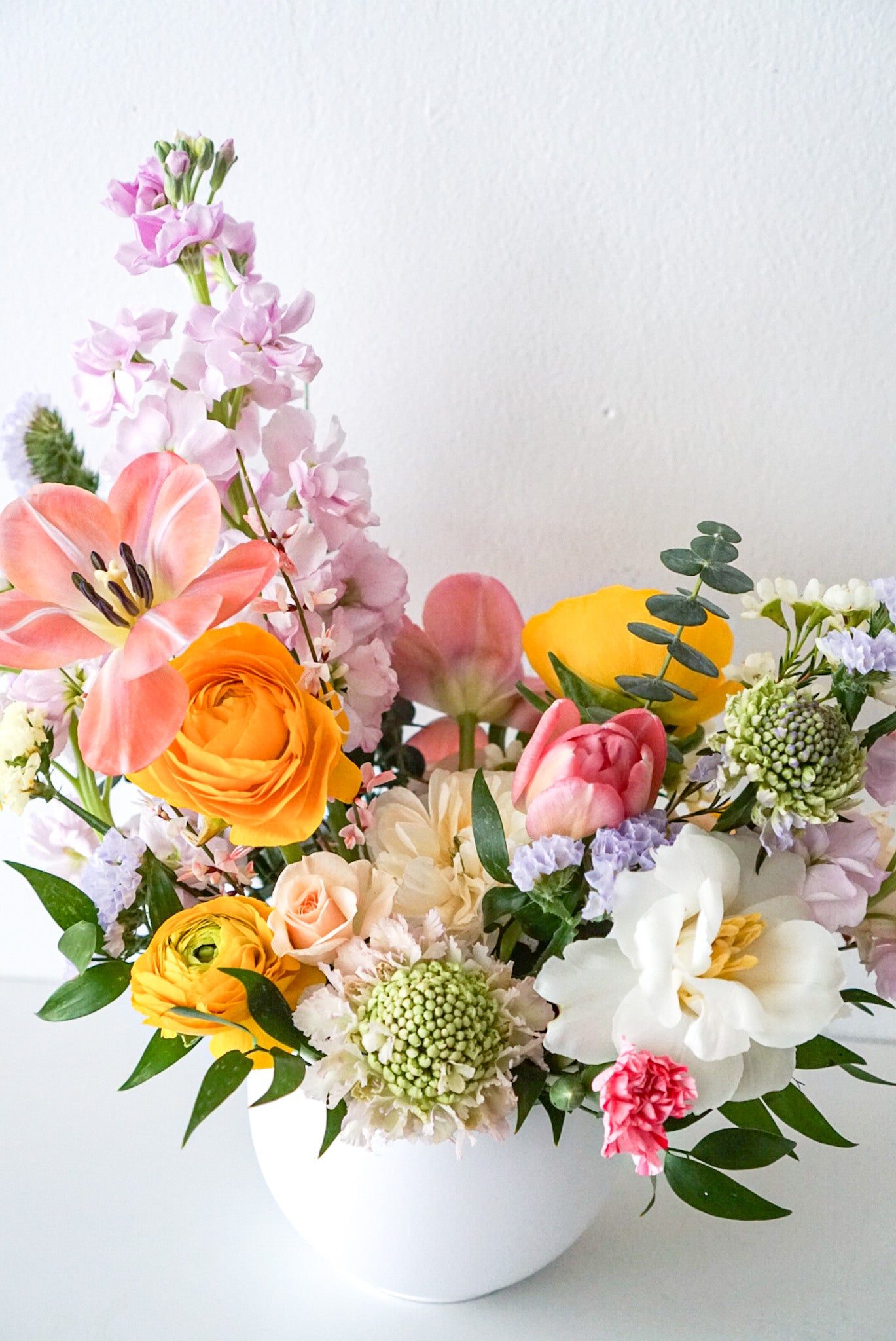 Like a spectacular spring sunrise, this radiant arrangement of yellow, peach, pink blooms, designed in a modern circular pot. This arrangement is sure to brighten anyone's day!  This arrangement features ranunculus, tulips, stocks, scabiosa, statics.