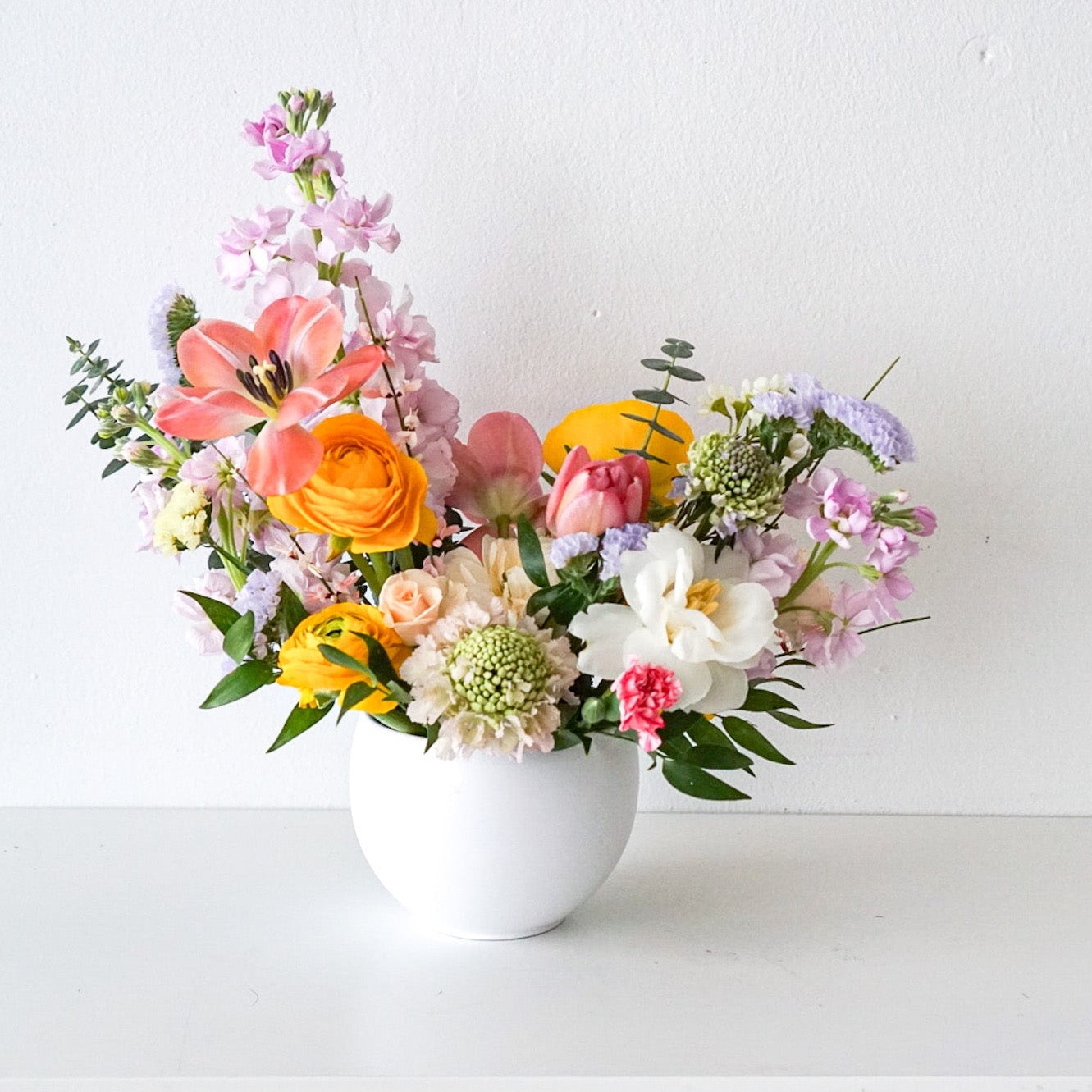 Like a spectacular spring sunrise, this radiant arrangement of yellow, peach, pink blooms, designed in a modern circular pot. This arrangement is sure to brighten anyone's day!  This arrangement features ranunculus, tulips, stocks, scabiosa, statics.