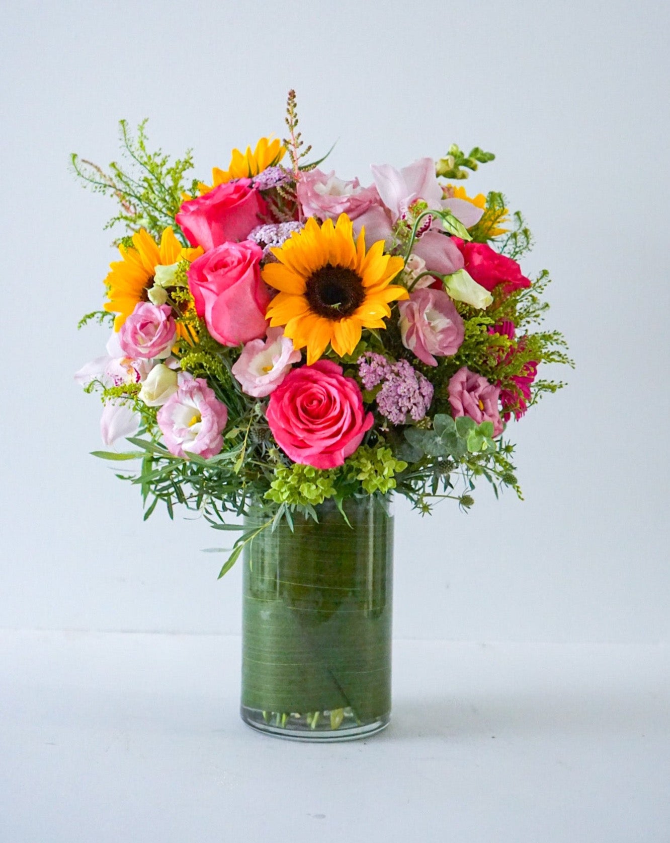  A bright summery arrangement, Summer Soiree is an ideal gift sure to brighten their day. This vivacious bouquet is an assortment of hot pink, yellow, orange and green summer blooms.  Delivered in a clear cylinder vase, lined with green ti leave Sunflowers, roses, lisianthus, yallow, cymbidium orchid, snap dragon, solidago, green hydrangeas. The Flower Nook- Toronto Florist- Flowers delivery Toronto