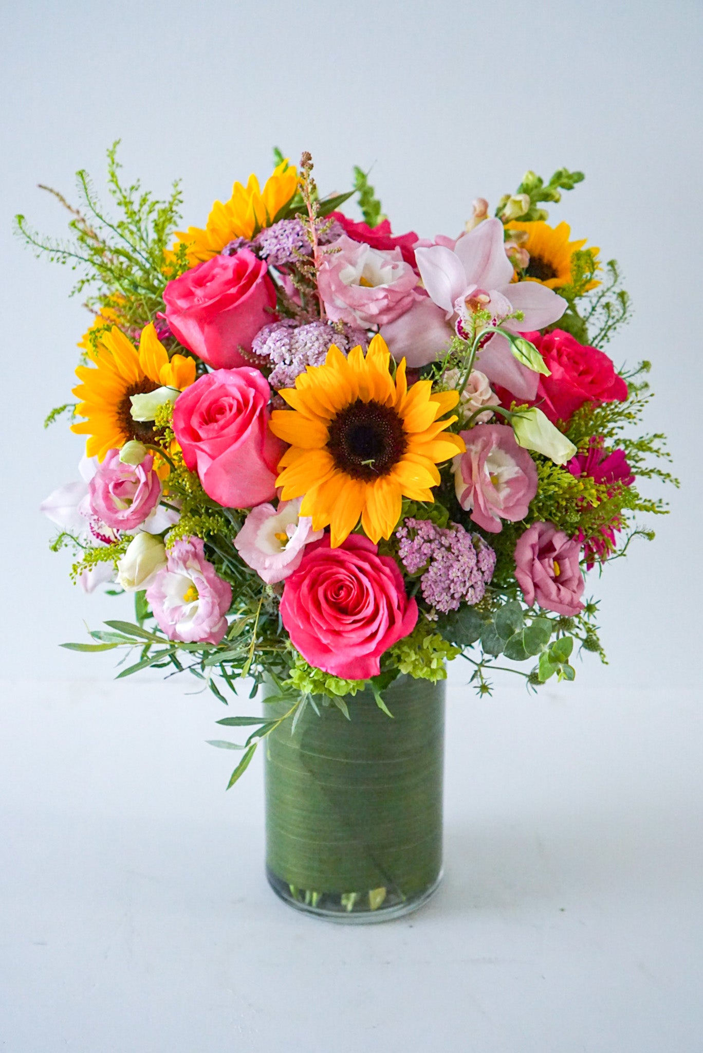  A bright summery arrangement, Summer Soiree is an ideal gift sure to brighten their day. This vivacious bouquet is an assortment of hot pink, yellow, orange and green summer blooms.  Delivered in a clear cylinder vase, lined with green ti leave Sunflowers, roses, lisianthus, yallow, cymbidium orchid, snap dragon, solidago, green hydrangeas. The Flower Nook- Toronto Florist- Flowers delivery Toronto