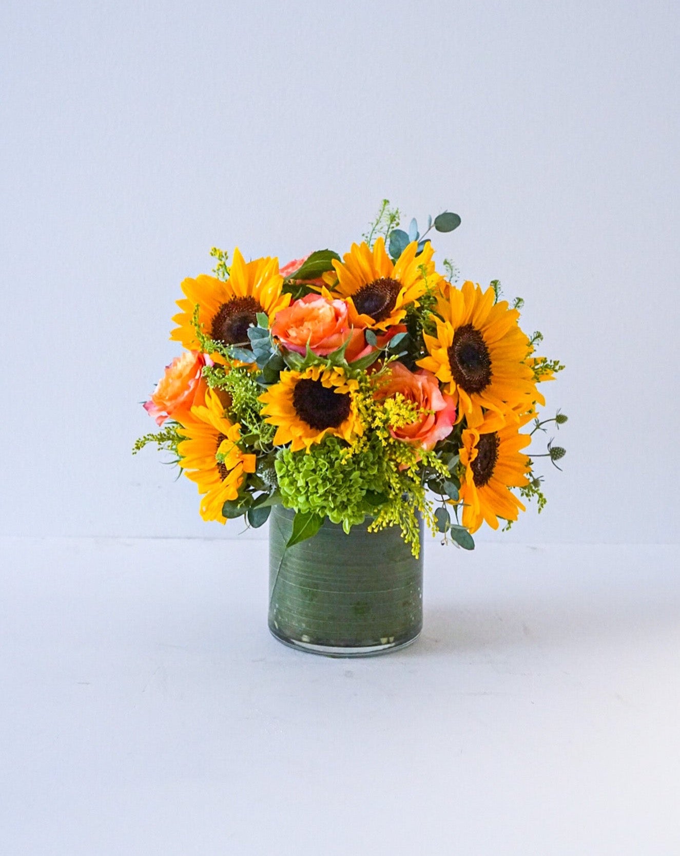 Brighten up a table, send get well wishes, or simply sprinkle sunshine on someone's day with this summer flower arrangement.  Local sunflowers steal the show in this simple arrangement. Also featured: green hydrangeas, free spirit roses, designed in the clear glass leaf-lined cylinder.